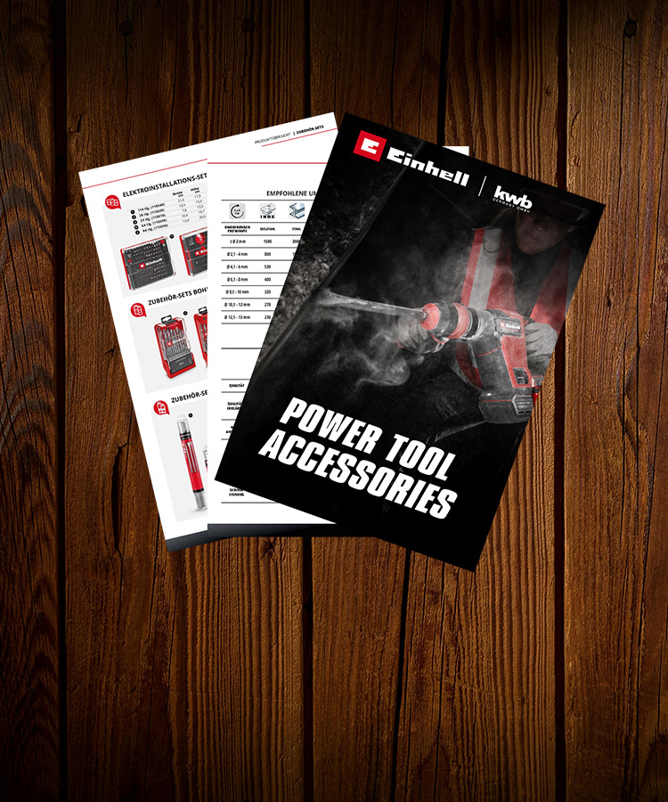 Einhell/kwb product catalogues placed on a dark wooden surface. The front catalogue shows a worker using a rotary hammer with the title ‘POWER TOOL ACCESSORIES’, while the one behind displays product overviews and accessory sets.