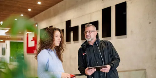 Two people are standing in the Einhell entrance area, engaged in conversation. A man is holding a tablet while a woman listens. The large Einhell logo is visible on the wall in the background.