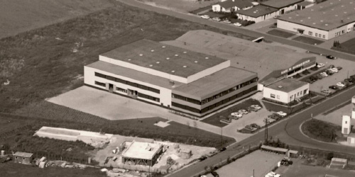 Historic aerial photograph of an industrial building with a large warehouse complex and surrounding parking areas. The site is located in a spacious industrial zone, surrounded by open land and several smaller facilities.