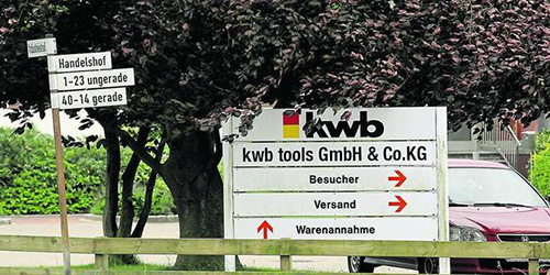 Company sign of kwb tools GmbH & Co. KG with directional information for visitors, shipping, and goods reception. Trees and a parked car are visible in the background, along with street signs for the ‘Handelshof’ area.